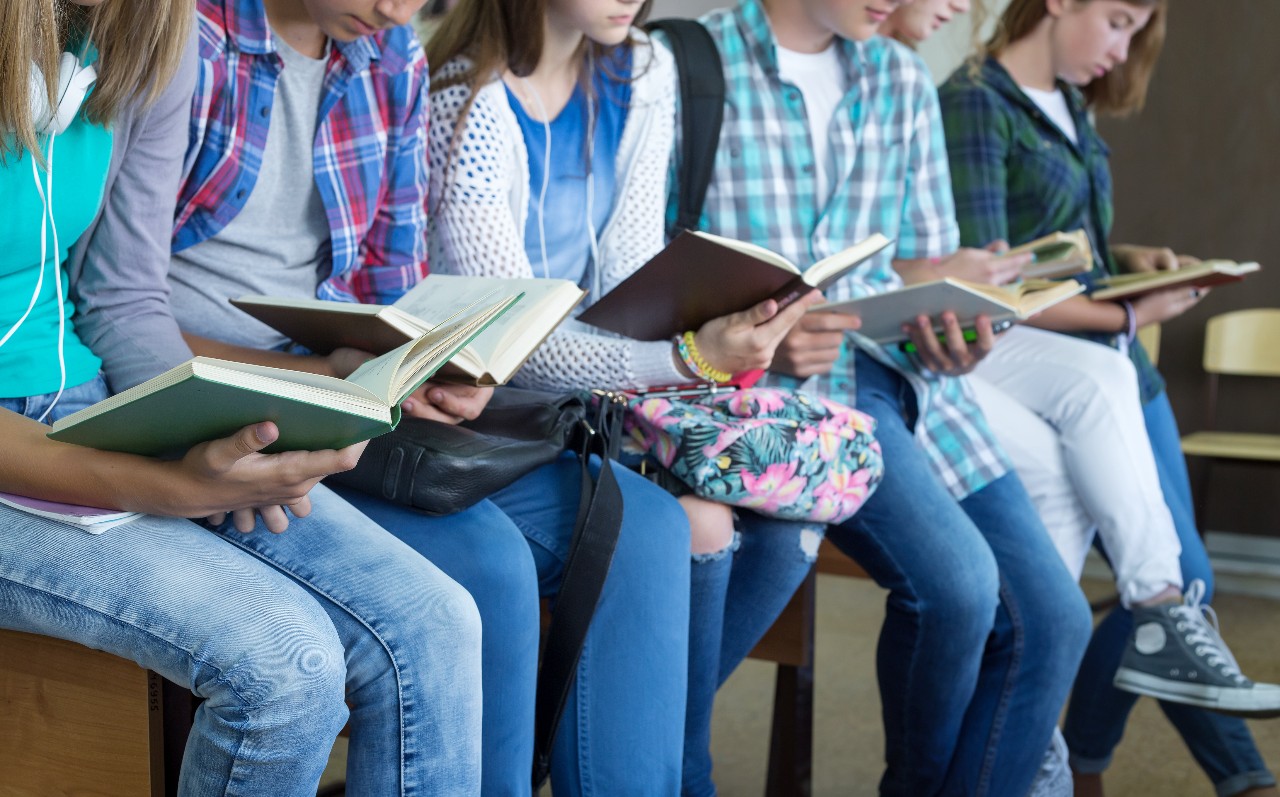 Students lined up on a bench reading books.